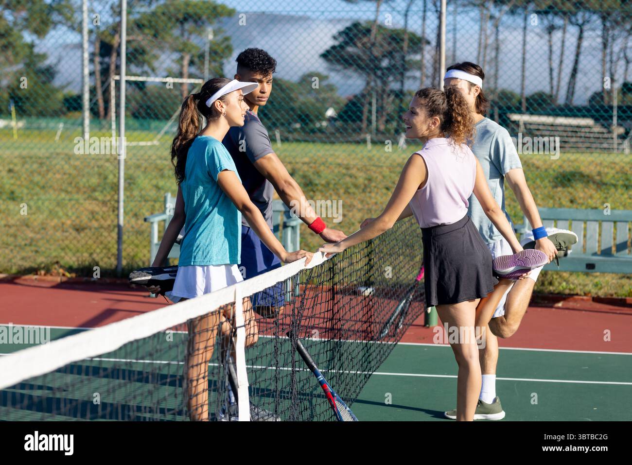 Divers joueurs de tennis en vêtements de sport penchés sur le filet sur le court du parc tenant des raquettes et bavardant Banque D'Images