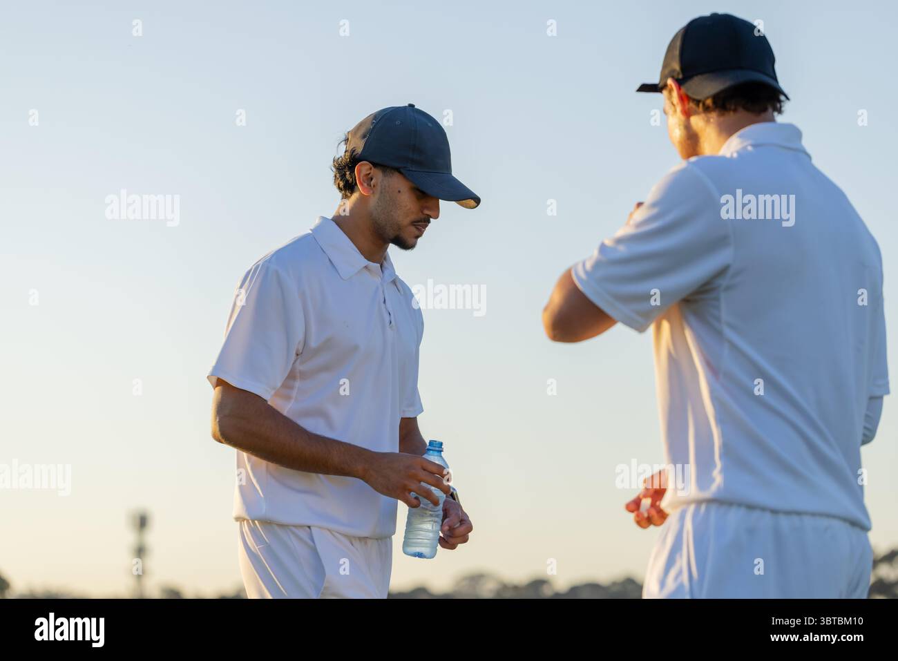 Divers coéquipiers masculins portant des polos blancs et des casquettes debout sur le terrain de cricket, tenant une bouteille d'eau Banque D'Images