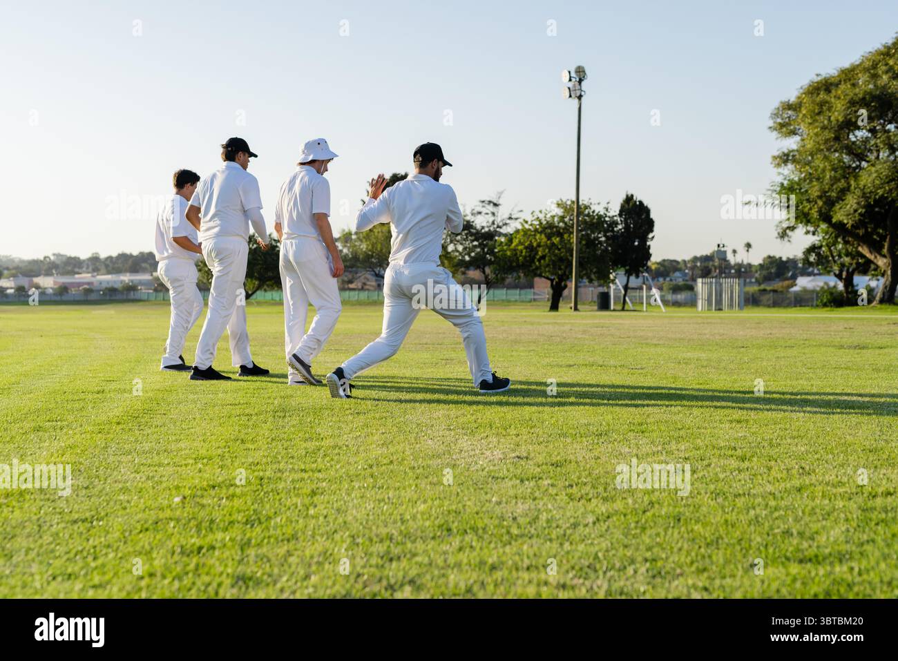 Divers coéquipiers masculins marchant en file unique à travers le terrain de cricket portant des kits blancs à la lumière du soleil Banque D'Images