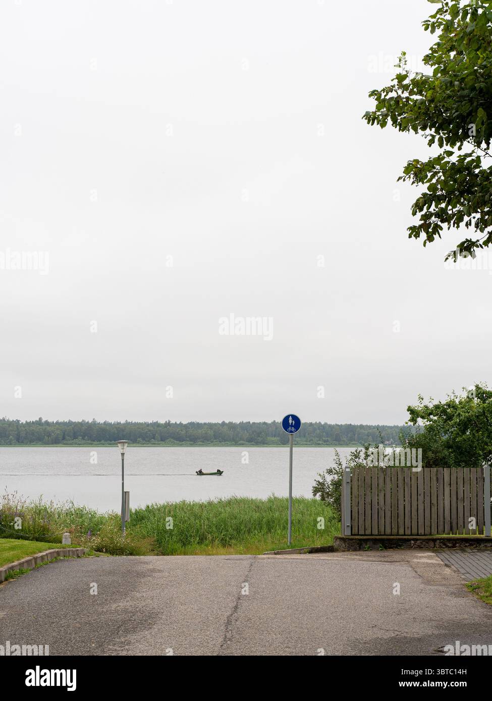 Chemin pavé menant à la rive du lac par une journée nuageuse en Estonie, avec panneau piétonnier et bateau lointain - atmosphère de campagne tranquille. Banque D'Images