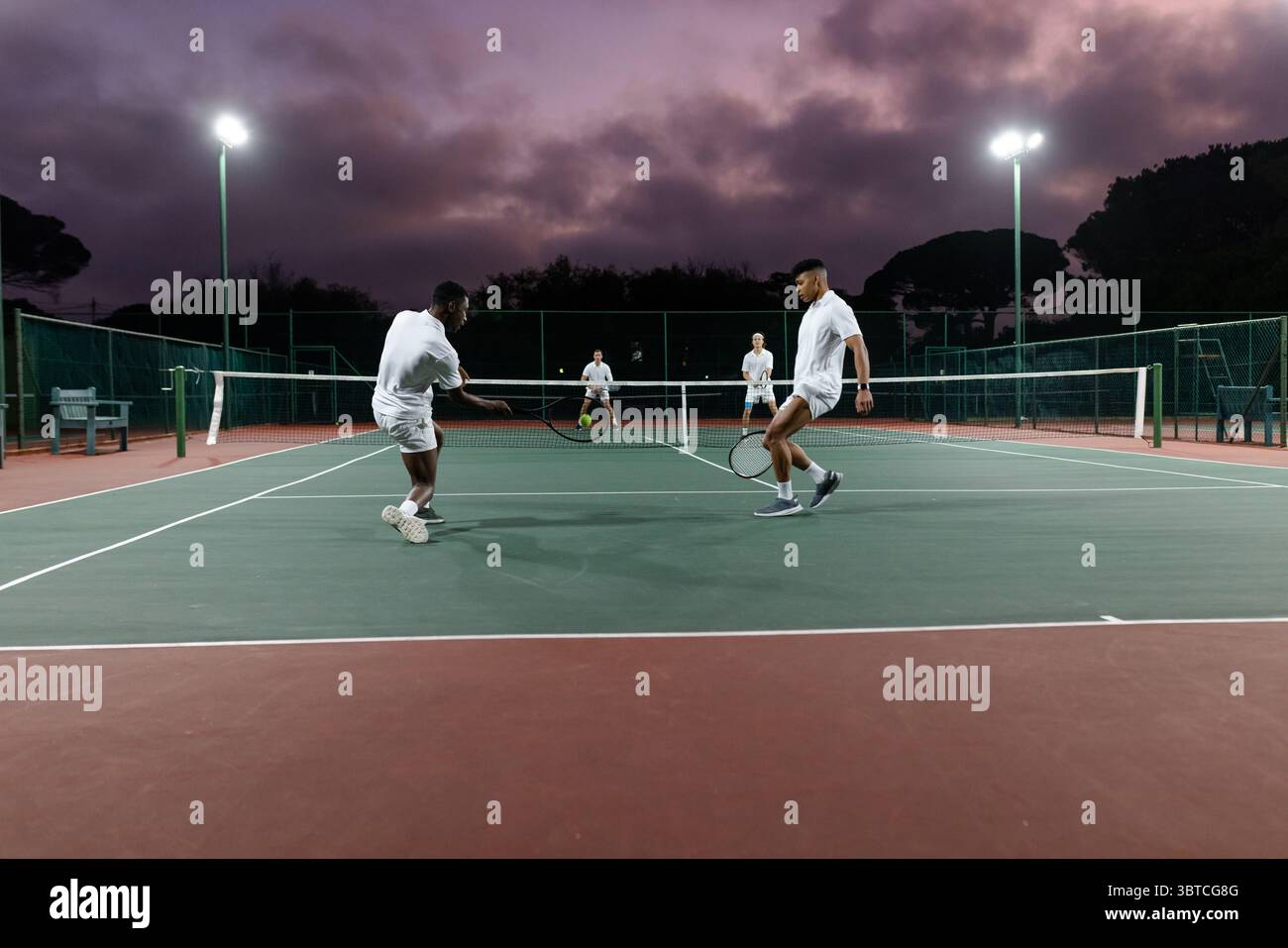 Divers joueurs de tennis masculins volants tenant des raquettes près du filet au crépuscule sous les projecteurs près de la clôture Banque D'Images