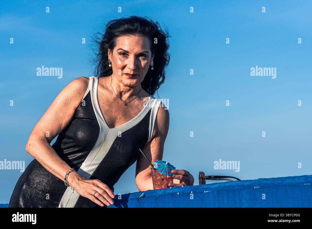 Femme senior élégante profitant d'un cocktail rafraîchissant sur un bar sur le toit, appuyée contre une balustrade bleue sous un ciel clair, rayonnant de confiance et de corps Banque D'Images