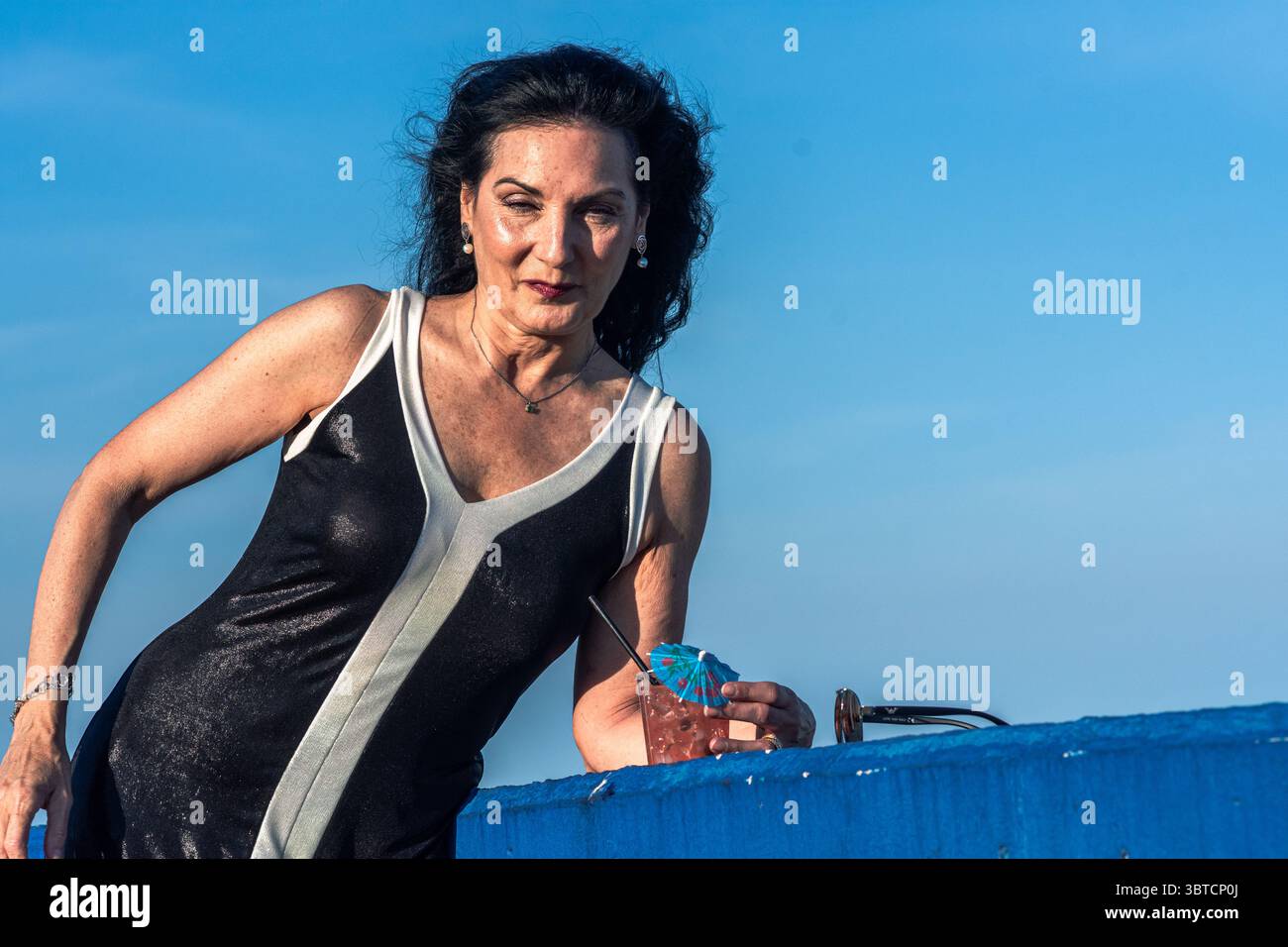 Modèle senior élégant avec les cheveux foncés et une élégante robe noire et blanche profite d'un cocktail rafraîchissant dans un ciel bleu vibrant, incarnant le positi corporel Banque D'Images Modèle senior élégant avec les cheveux foncés et une élégante robe noire et blanche profite d'un cocktail rafraîchissant dans un ciel bleu vibrant, incarnant le positi corporel Banque D'Images