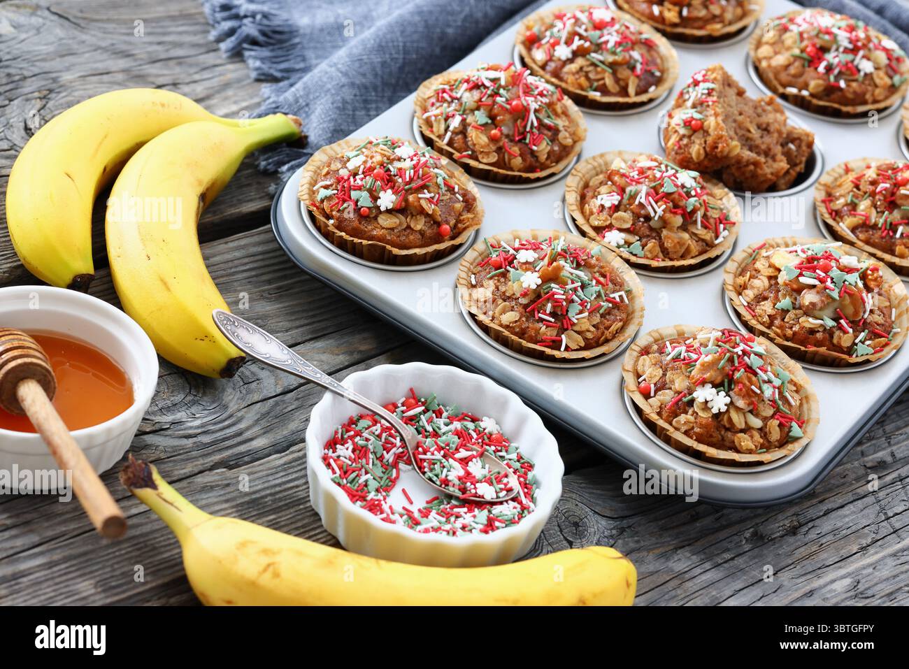 muffins au pain à la cannelle et à la banane recouverts de saupoudrés de noël dans un moule à muffins sur une table en bois rustique avec du miel et des ingrédients, plat, horizonta Banque D'Images