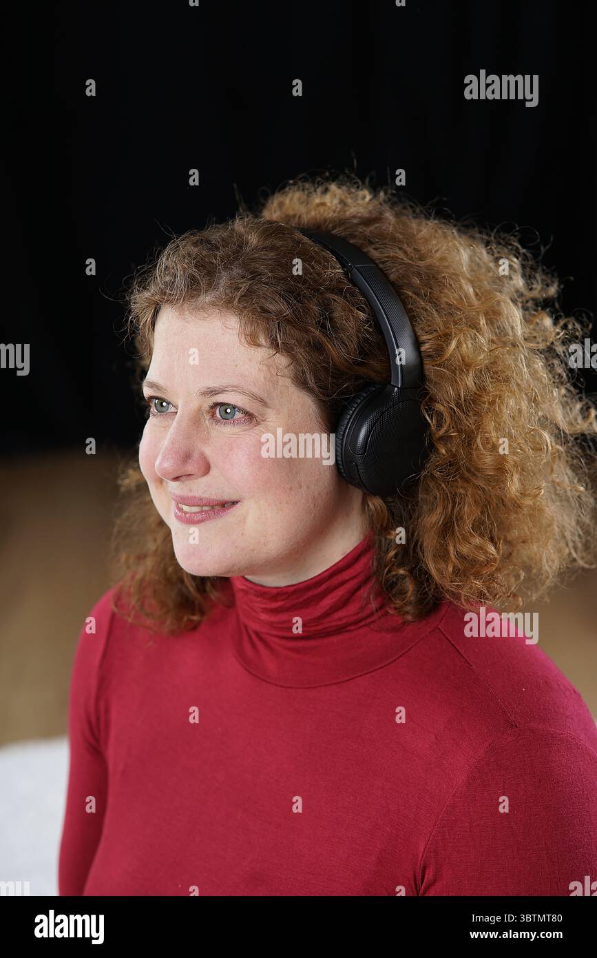 Femme en pull à col roulé rouge avec des écouteurs noirs appréciant la musique, les yeux ouverts, souriant Banque D'Images