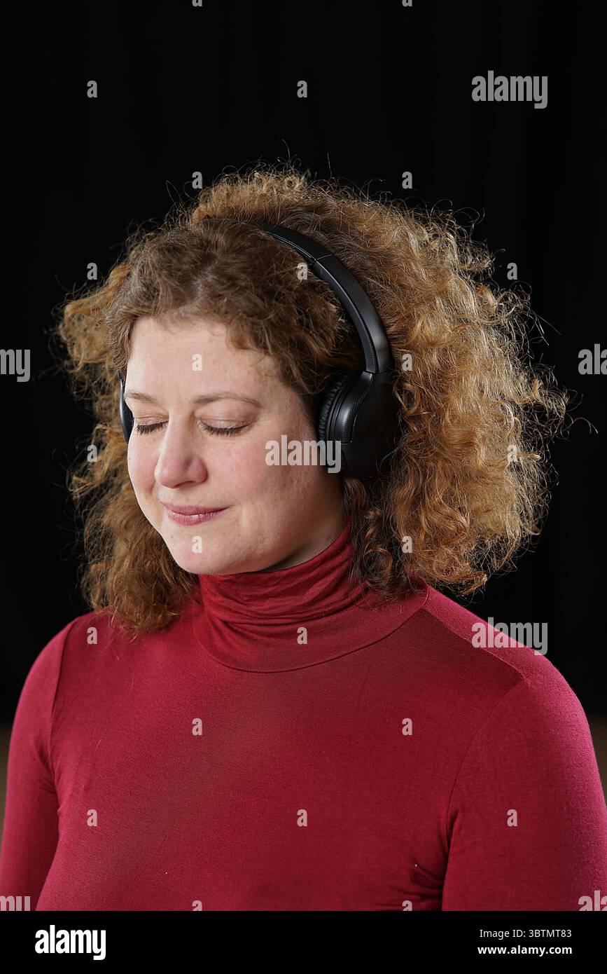 Femme en pull à col roulé rouge avec des écouteurs noirs appréciant la musique, les yeux fermés Banque D'Images