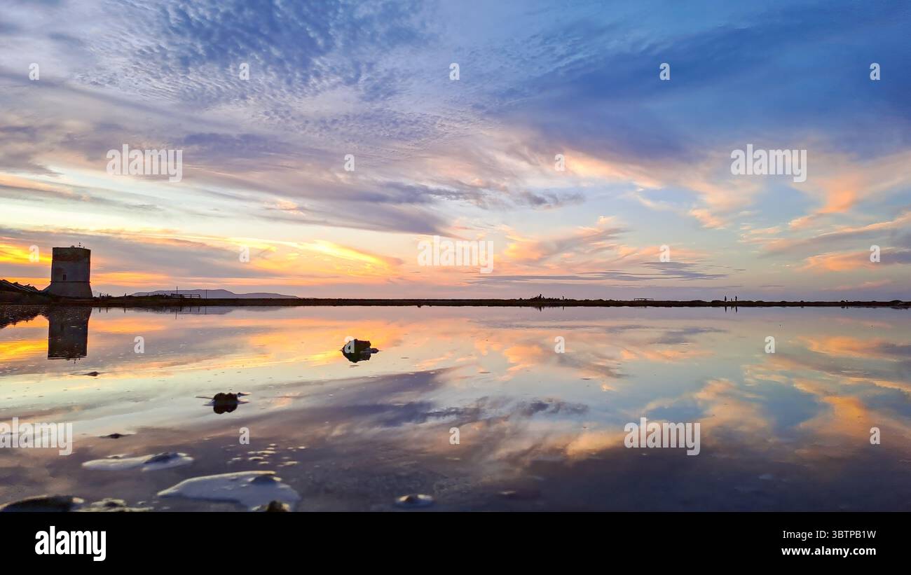 Ciel coloré de coucher de soleil reflété dans les plaines de sel encore près de Paceco, Trapani, Sicile, avec un vieux moulin à vent visible à l'horizon. Banque D'Images