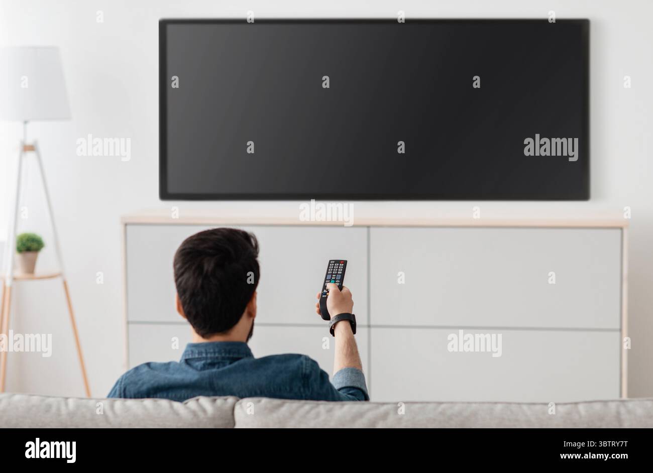 Homme regardant la télévision, allumant le téléviseur à écran plat plasma, pointant la télécommande vers un écran de télévision vide sur un mur blanc Banque D'Images