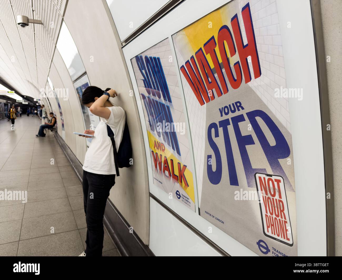 Une femme passager vérifie le téléphone pour les heures de train contre le mur de plate-forme tubulaire avec l'affiche « Watch Your Step Not Your Phone » dans le métro londonien Banque D'Images