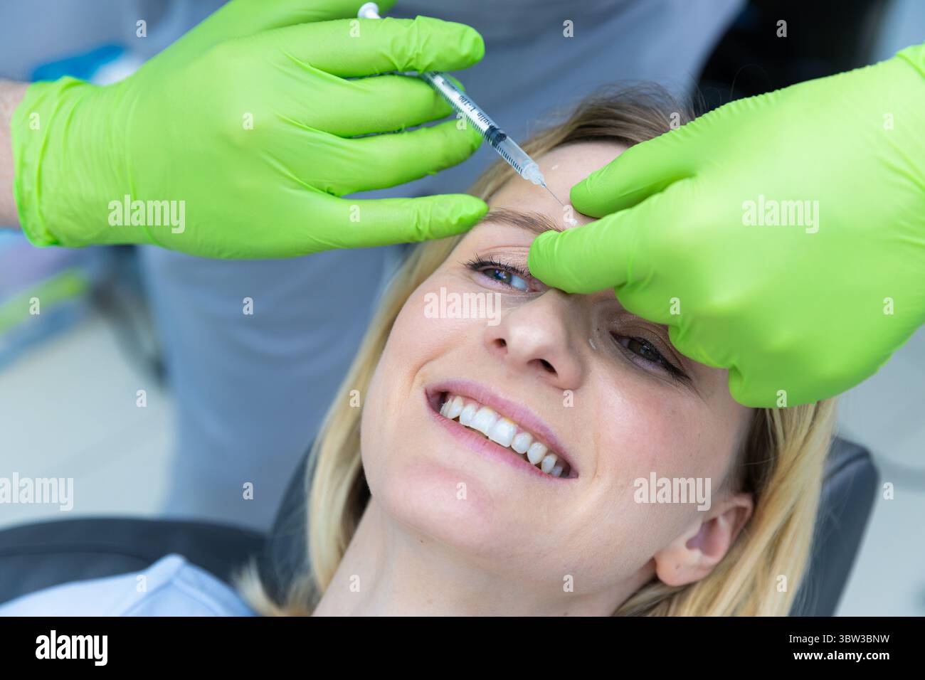 Une procédure esthétique professionnelle : gros plan d'une femme souriante recevant une injection de Botox dans une clinique moderne avec des gants verts et confortables Banque D'Images