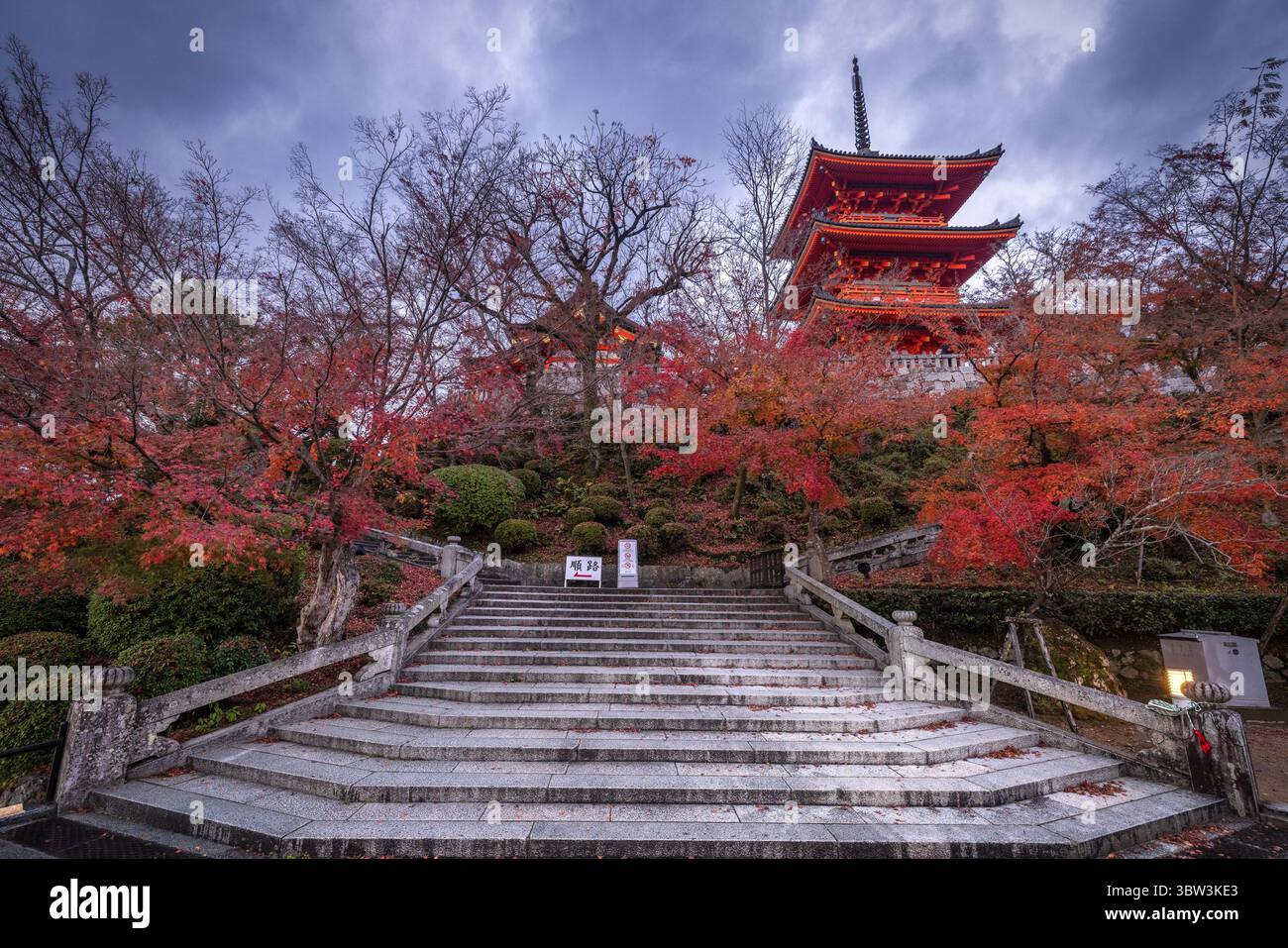 Vue d'une pagode rouge vibrante nichée au sommet d'une colline, encadrée par un feuillage d'automne ardent et un escalier en pierre sous un ciel nuageux, Kyoto, Kyoto, Japon. Banque D'Images