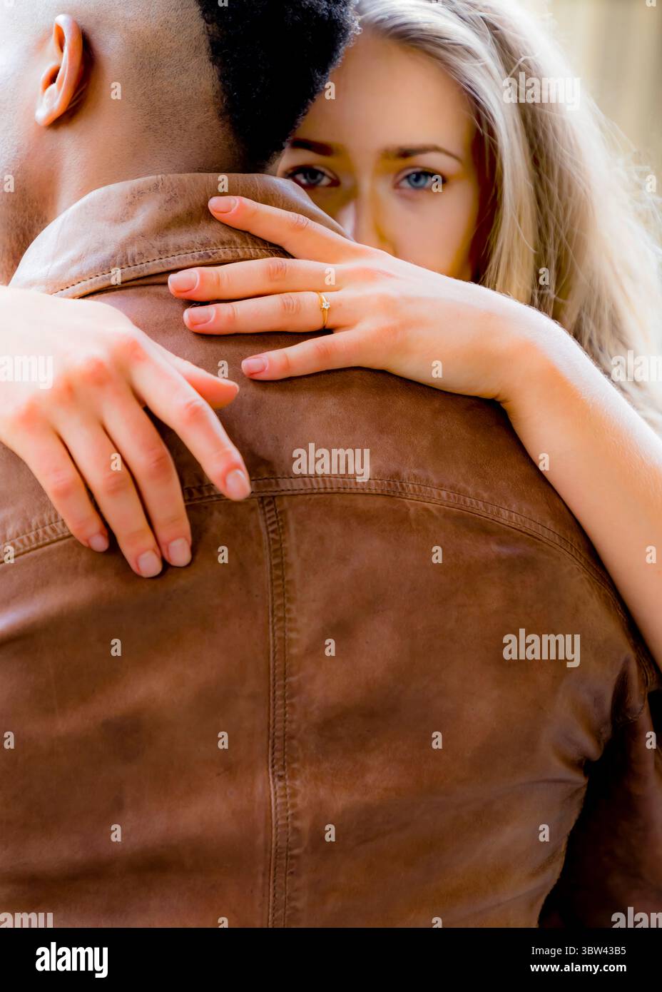 Une femme aux cheveux blonds et aux yeux bleus embrasse un homme par derrière. Elle porte une bague au doigt. L'homme porte une veste en cuir marron. Banque D'Images