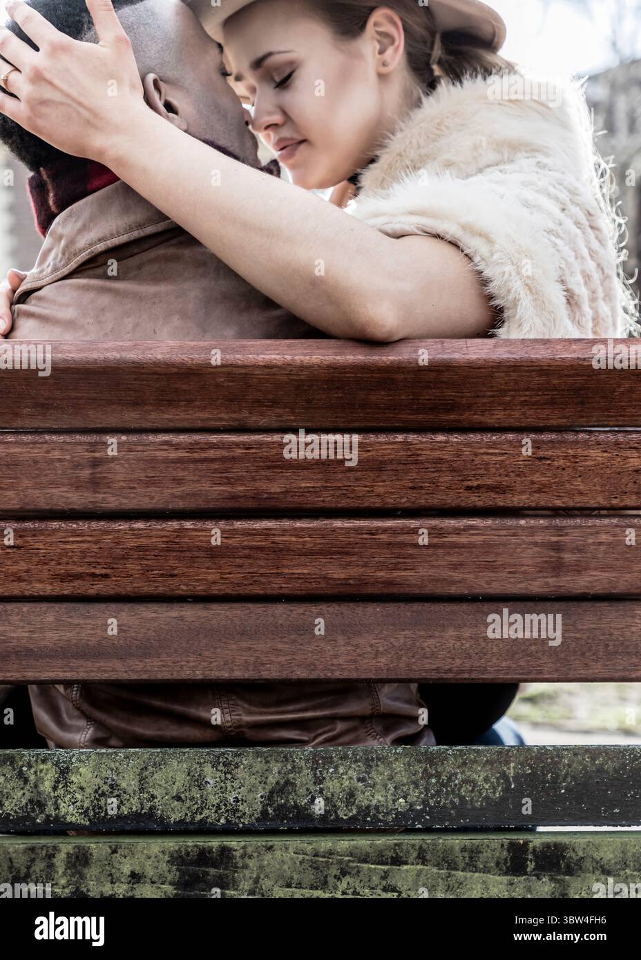 Un couple s'embrasse sur un banc. La femme porte un chapeau et une étole de fourrure, et elle tient la tête de l'homme. L'homme porte une veste en cuir. Banque D'Images