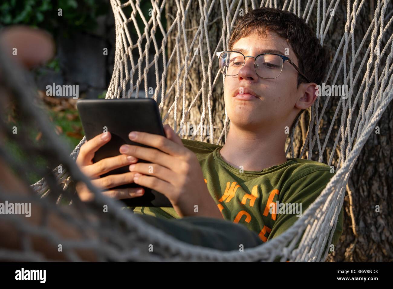 Un jeune homme se relaxant dans un hamac à l'extérieur est concentré sur la lecture d'un e-livre sur un e-lecteur moderne, représentant un style de vie numérique, e-learning, et lei Banque D'Images