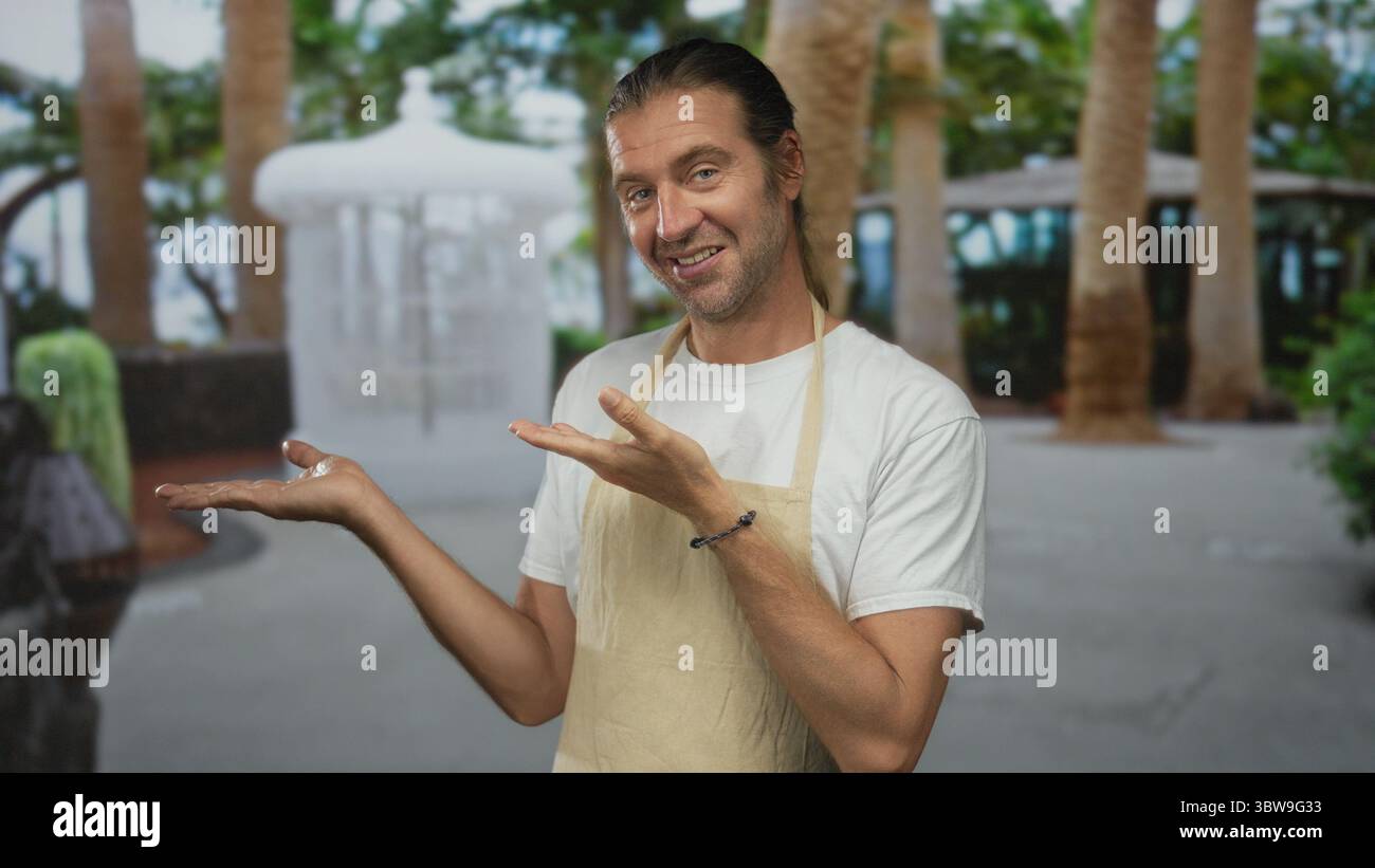 Blond Man in Apron montre des palmiers ouverts sur fond de forêt luxuriante tout en souriant chaleureusement ; bienvenue. Banque D'Images Blond Man in Apron montre des palmiers ouverts sur fond de forêt luxuriante tout en souriant chaleureusement ; bienvenue. Banque D'Images