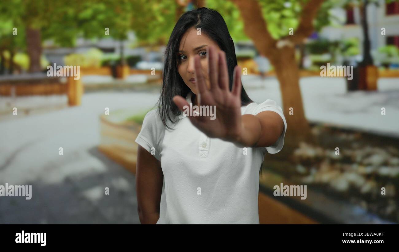 Jeune femme à l'extérieur dans un parc avec arrêt de signalisation à main prolongée, entourée d'arbres verts vibrants et de fond urbain, exprimant la confiance et comme Banque D'Images