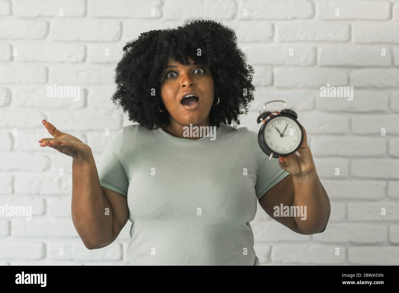 Femme afro-américaine surprise tenant le réveil dans une main sur fond de mur de briques. Concept de délai et de temps Banque D'Images