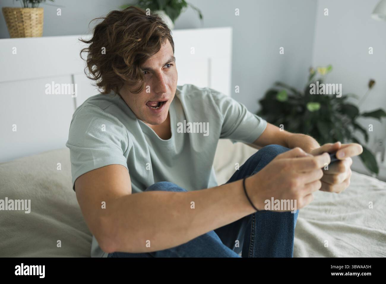 Huebscher aufgeregter Gamer, der auf einem Bett sitzt und Videospiele auf einer Konsole spielt. Wireless-Controller und Online-Spiel-Technologien Konz Banque D'Images