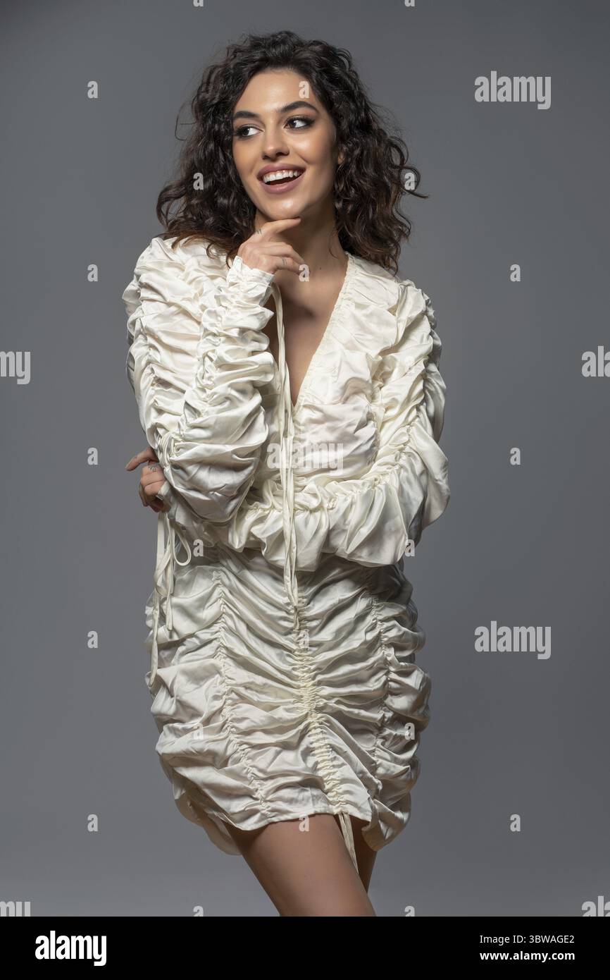 Jeune femme mince attrayante et joyeuse avec des cheveux foncés ondulés portant une robe de fête courte drapée blanche posant sur fond de studio gris, riant à la recherche Banque D'Images