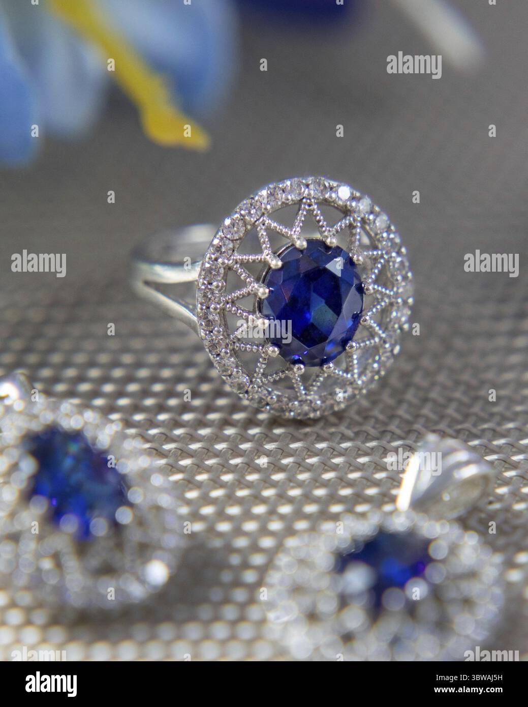 Bague en argent avec une grande pierre précieuse bleue et de petits diamants autour d'elle entourée d'autres bijoux sur une surface grise Banque D'Images