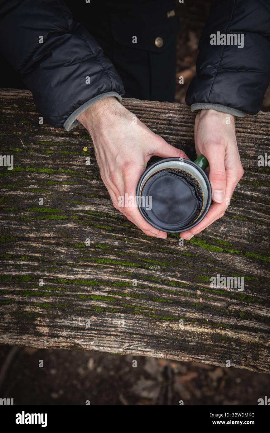 Vue de haut en bas des mains tenant une tasse de café noir sur une table en bois humide altérée, une personne portant une veste en duvet, un cadre extérieur automne ou hiver. Banque D'Images