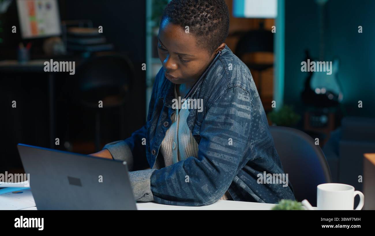 Télétravailleur afro-américain recevant un appel téléphonique d'un collègue de travail tout en travaillant à domicile sur un ordinateur portable. Freelance parlant avec un collègue à l'aide d'un smartphone tout en résolvant des tâches, caméra A. Banque D'Images