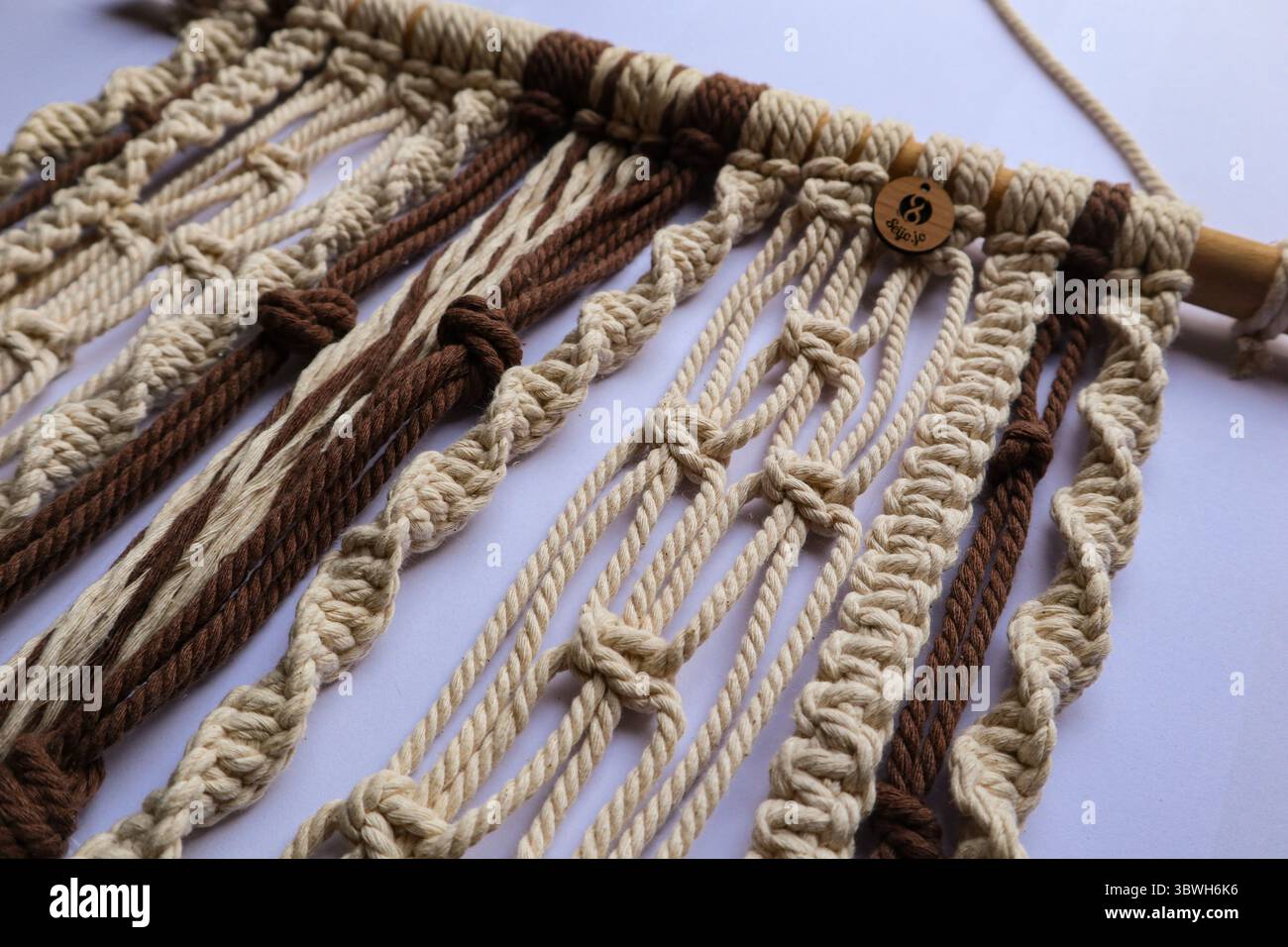 Gros plan d'une suspension murale en macramé aux tons neutres avec des nœuds complexes et une cheville en bois, sur fond de bois sombre Banque D'Images