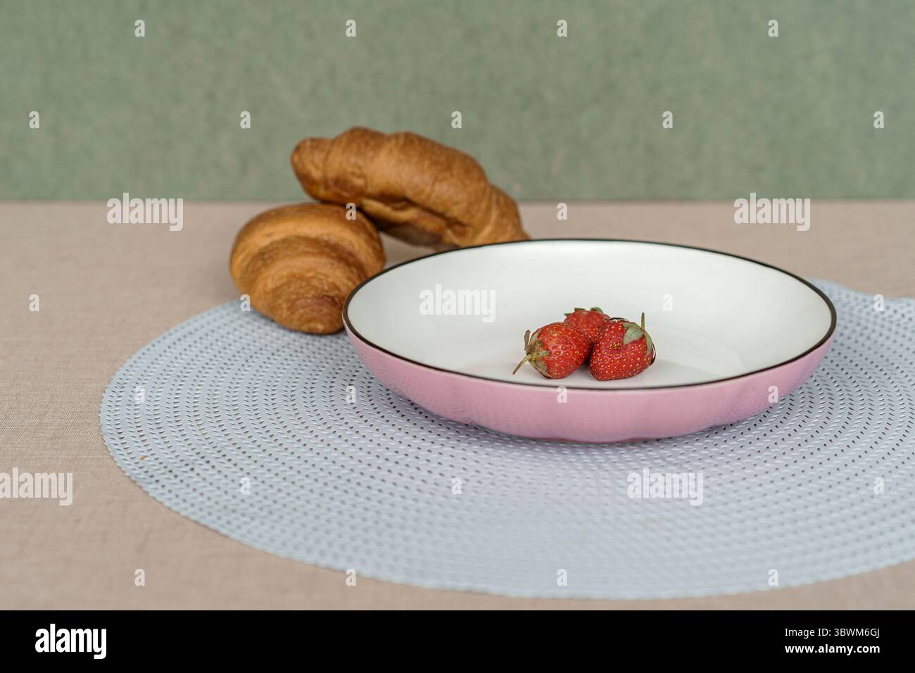 Petit déjeuner élégant encore de la vie. Fraises fraîches sur une plaque de céramique rose avec des croissants sur un napperon bleu et fond vert. Banque D'Images