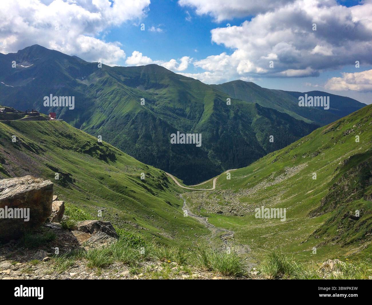 Vallée de montagne verte avec route sinueuse et pics spectaculaires dans les montagnes de Fagaras, Roumanie, sous un ciel partiellement nuageux. Banque D'Images