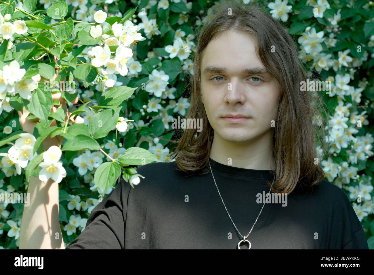 Un jeune homme aux cheveux longs pose près de fleurs blanches parfumées par une journée ensoleillée. Banque D'Images