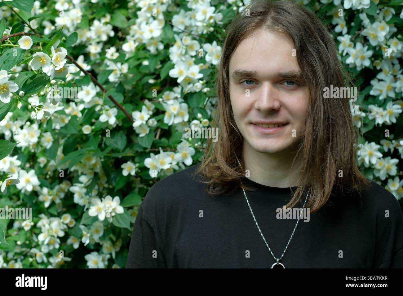 Un jeune homme aux longs cheveux bruns se tient souriant dans un jardin plein de fleurs. Banque D'Images