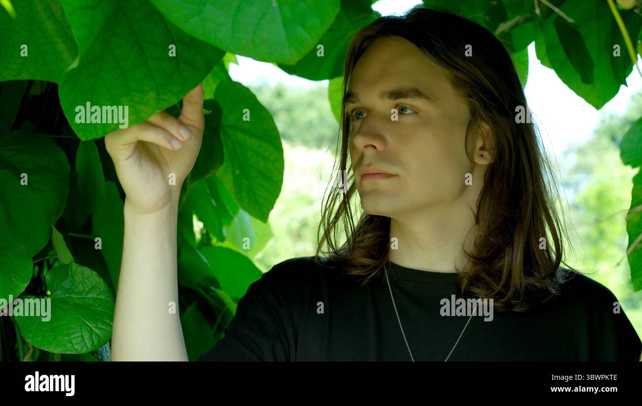 Un jeune homme touche doucement les feuilles dans un jardin tranquille, appréciant la beauté de la nature. Banque D'Images