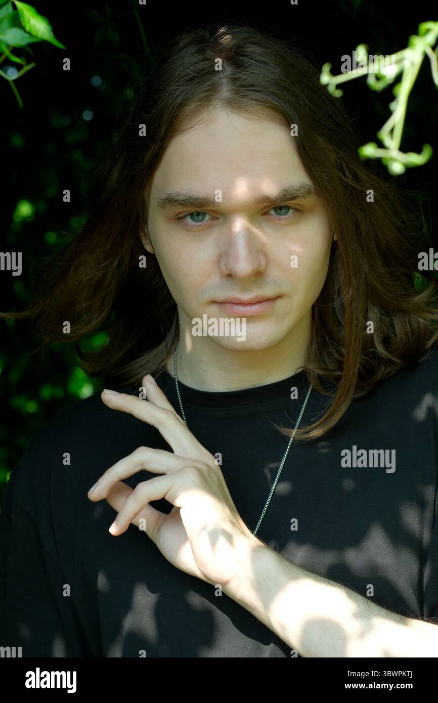 Un jeune homme aux cheveux longs se tient au milieu d'une verdure luxuriante, les ombres dansant sur son visage. Banque D'Images