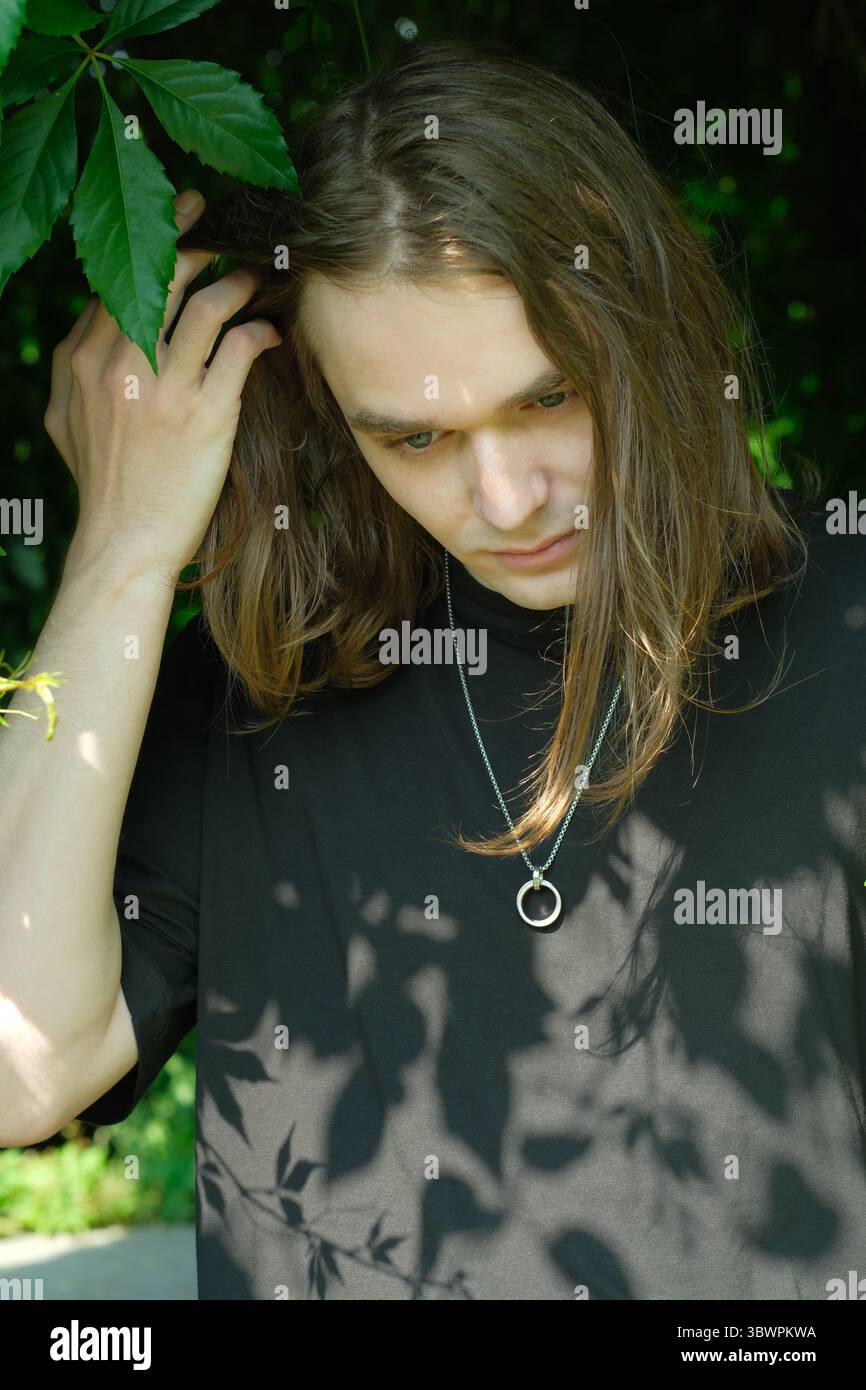 Un jeune homme se tient sous des branches feuillues, perdu dans la pensée et la sérénité. Banque D'Images