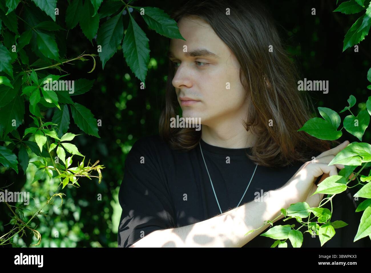 Un jeune homme aux cheveux longs regarde à travers un feuillage vert éclatant, captivé par la nature. Banque D'Images