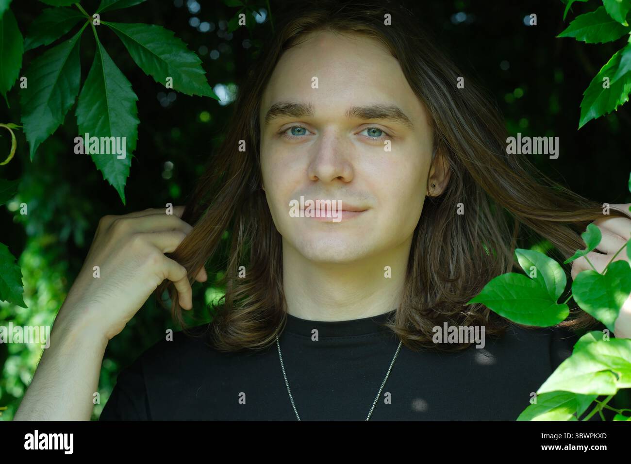 Un jeune homme sourit tout en tenant doucement ses longs cheveux dans un cadre vert éclatant. Banque D'Images
