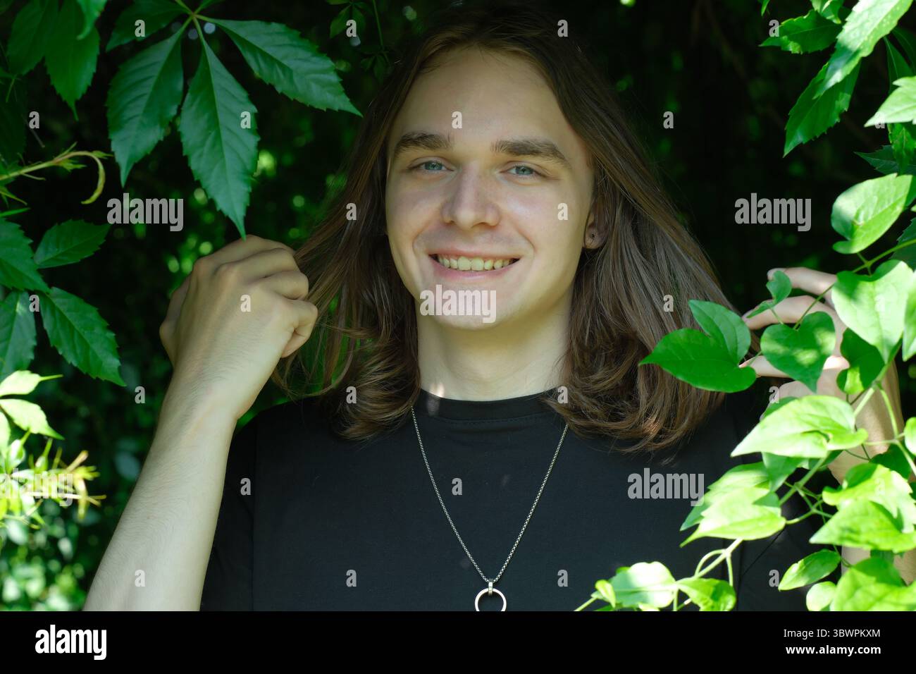 Un jeune homme joyeux touche ludique ses cheveux au milieu de feuilles vertes éclatantes. Banque D'Images