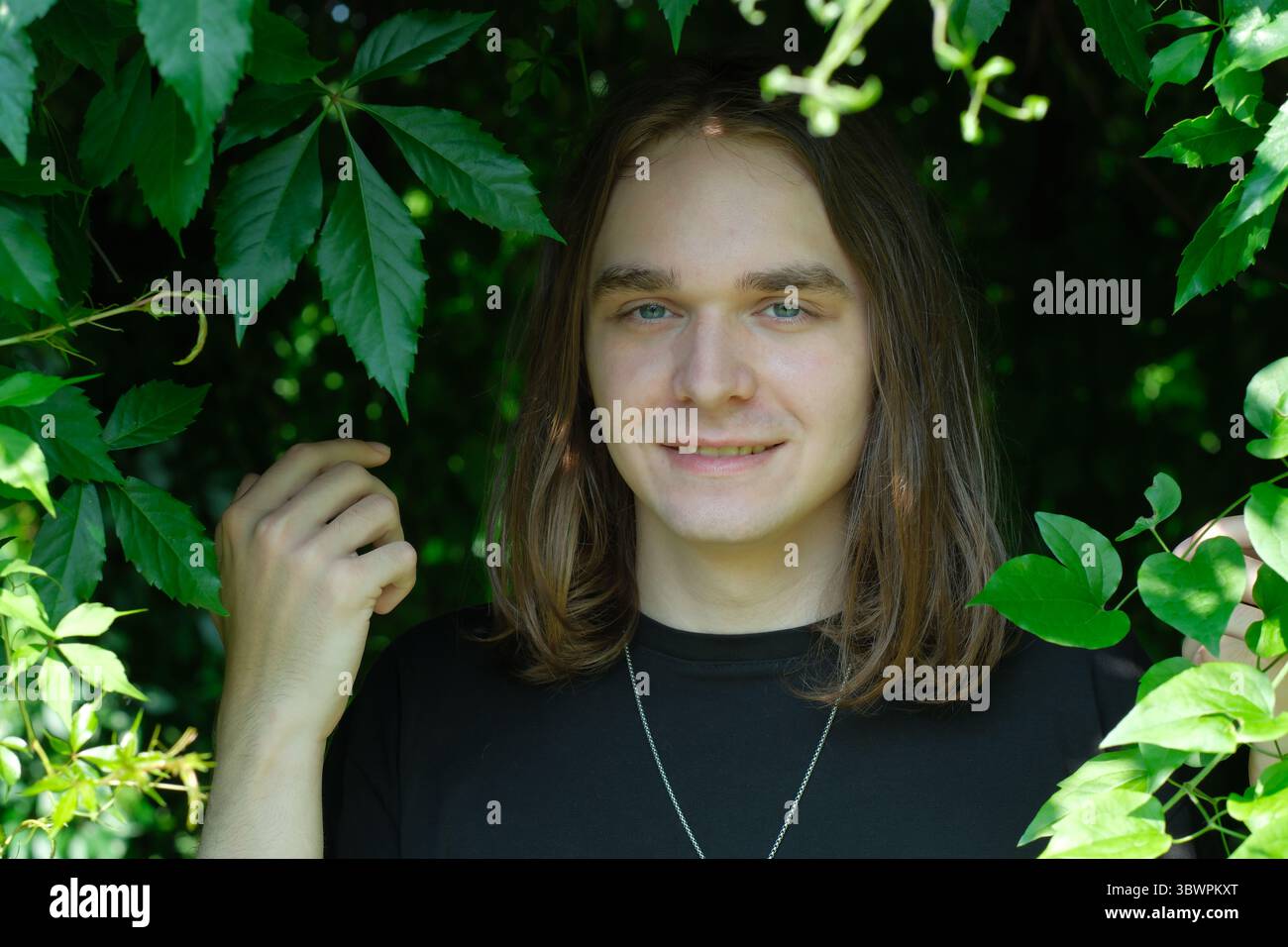 Un jeune homme se tient entouré de feuilles vertes vibrantes, souriant brillamment dans la nature. Banque D'Images