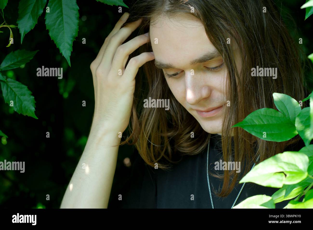 Un jeune homme aux cheveux longs se reflète parmi les feuilles vertes vibrantes, rayonnant de tranquillité. Banque D'Images