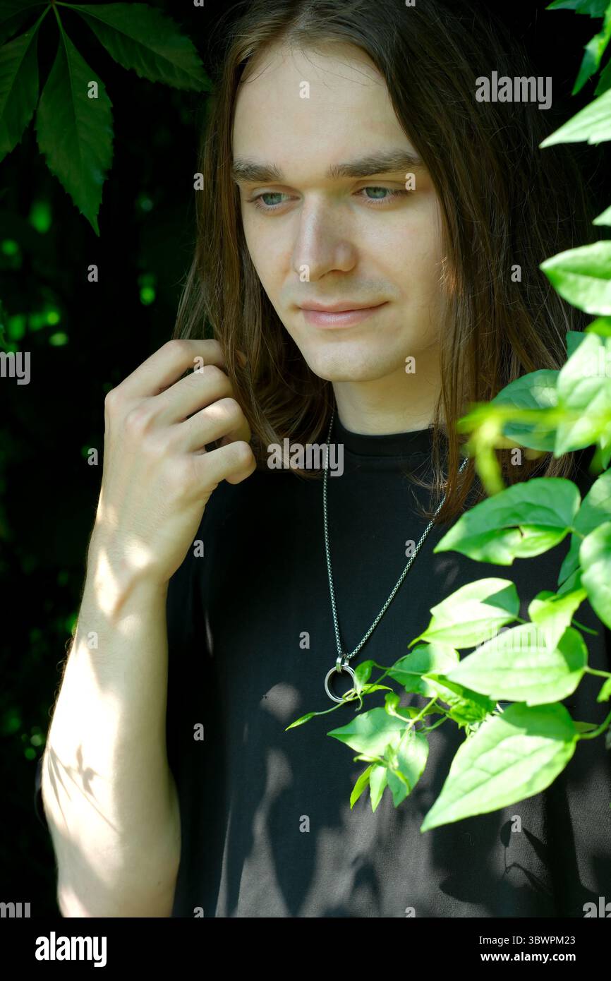 Un jeune homme se tient tranquillement parmi les feuilles vertes, la lumière du soleil illuminant son visage. Banque D'Images