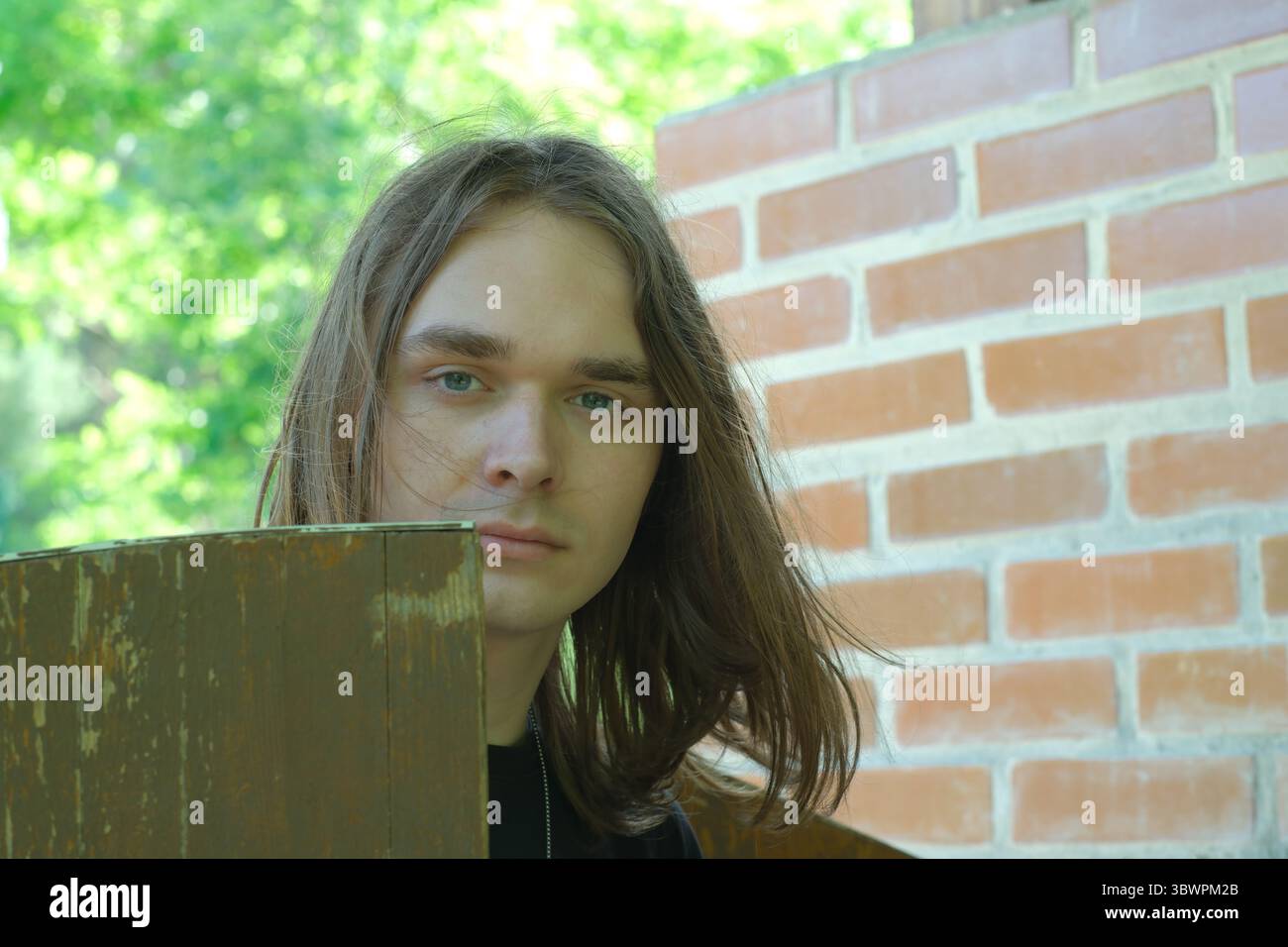 Un jeune homme avec des cheveux coulants se tient soigneusement à côté d'une clôture en bois rustique. Banque D'Images