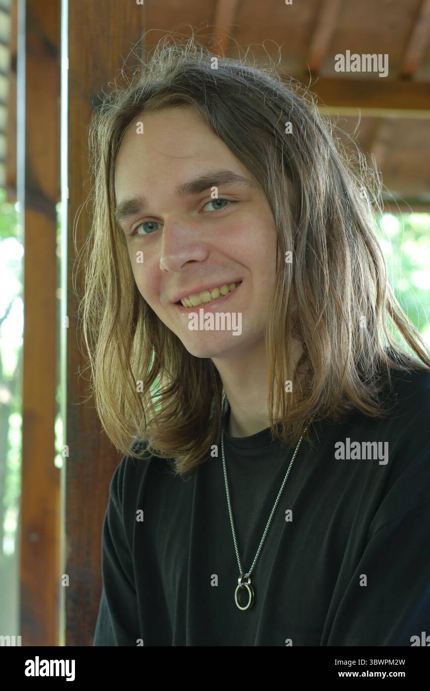 Jeune homme aux cheveux longs sourit chaleureusement tout en se relaxant dans un environnement naturel. Banque D'Images