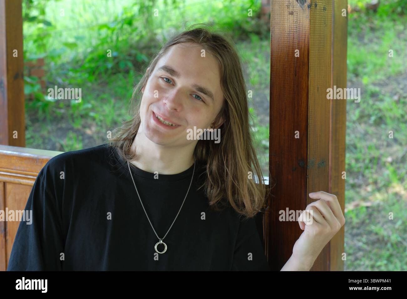 Un jeune homme aux cheveux longs sourit par un poteau en bois dans un cadre verdoyant. Banque D'Images
