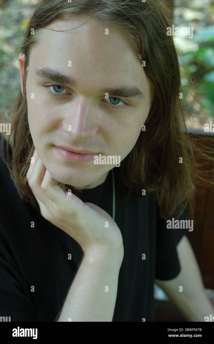 Un jeune homme aux cheveux fluides et aux yeux bleus frappants se détend dans un cadre extérieur serein. Banque D'Images