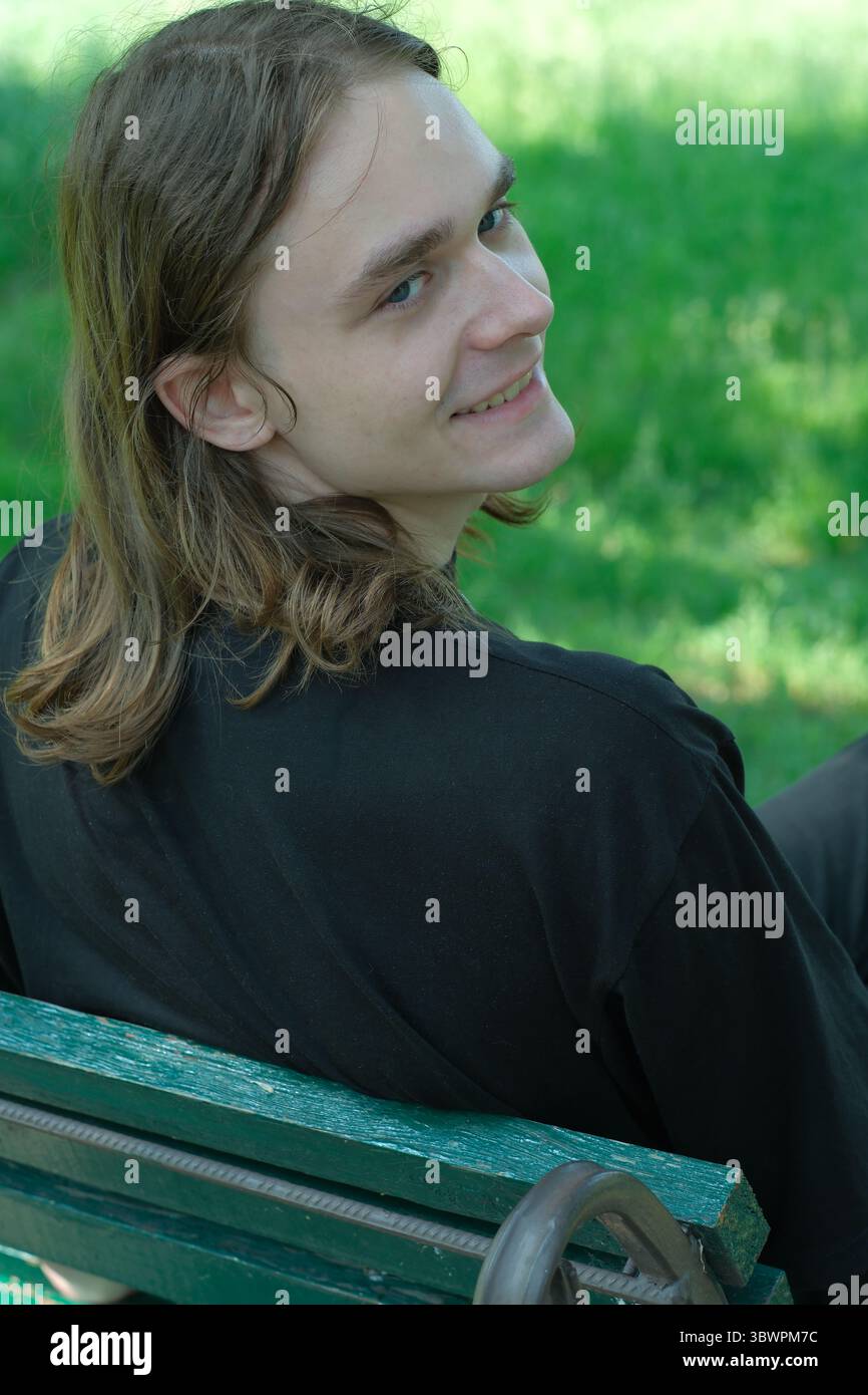 Jeune homme aux cheveux longs se détend sur un banc sous des arbres verts, souriant chaleureusement. Banque D'Images