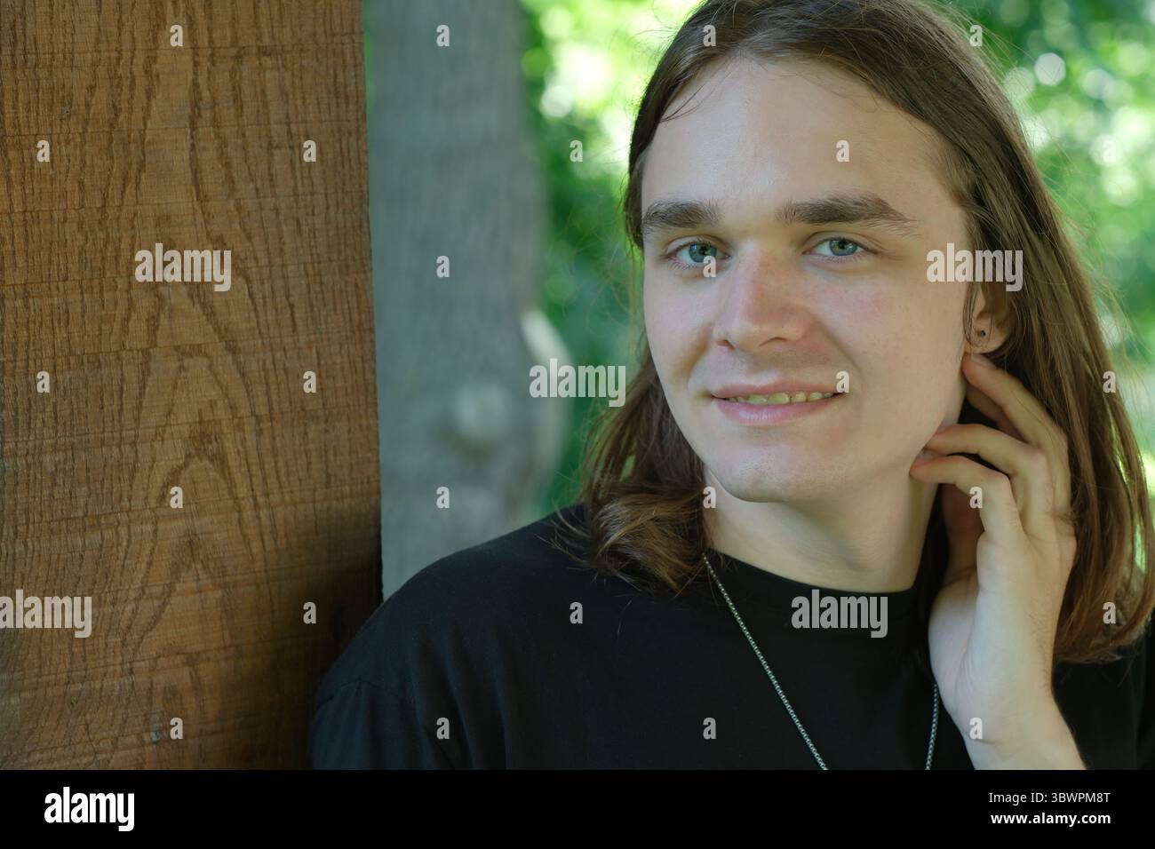 Un jeune homme confiant avec de longs cheveux pose près de la structure en bois, exsudant la chaleur. Banque D'Images