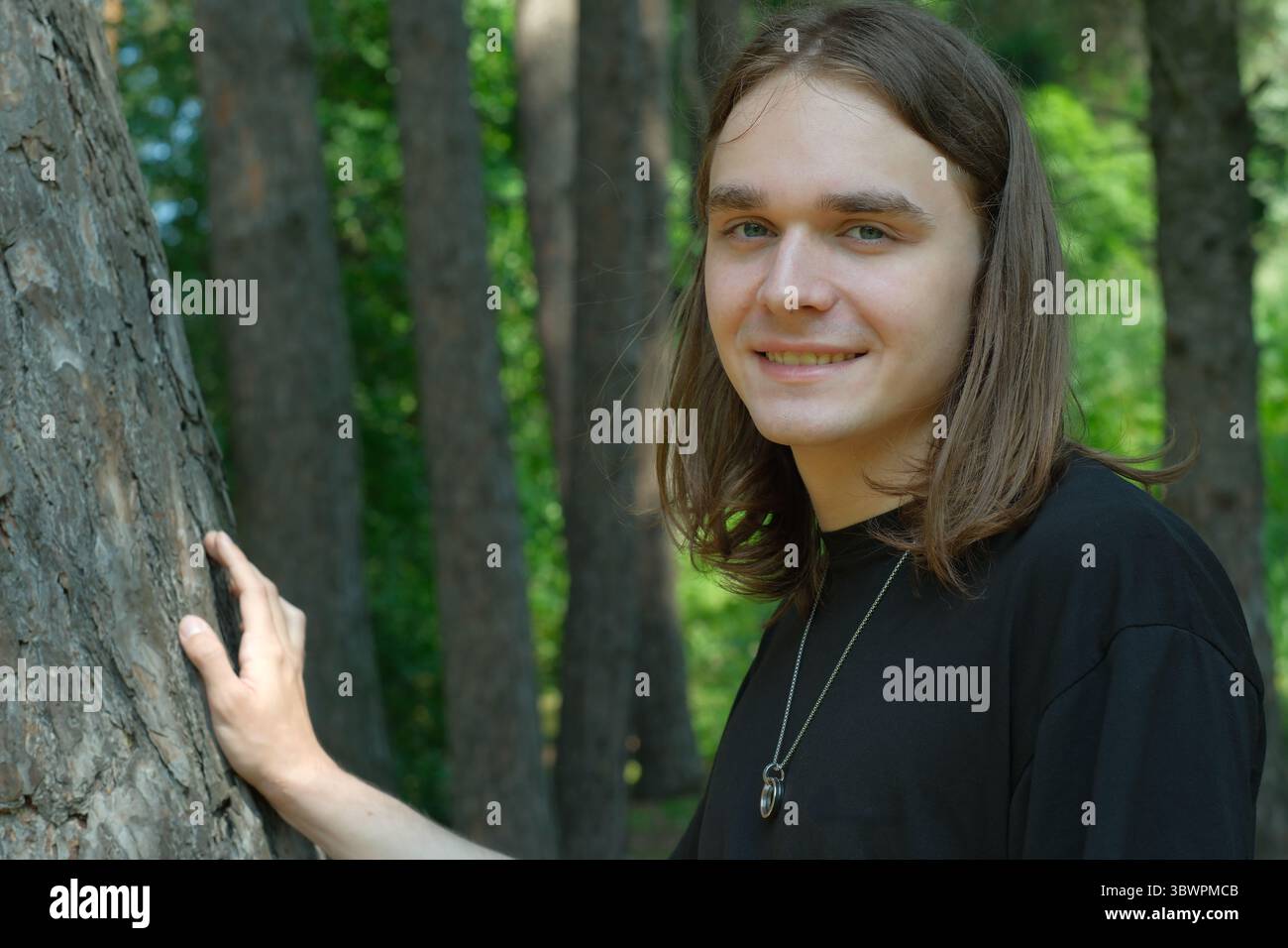 Un jeune homme se tient contre un arbre ancien, souriant au milieu d'arbres verdoyants. Banque D'Images