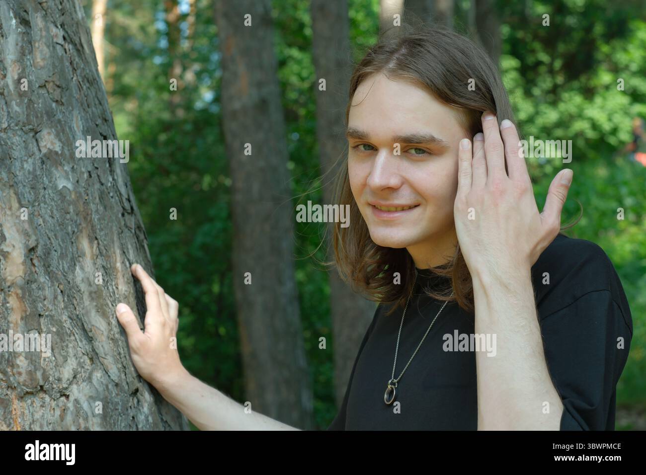 Un jeune homme se tient à côté d'un arbre, souriant doucement dans une forêt sereine en toile de fond. Banque D'Images