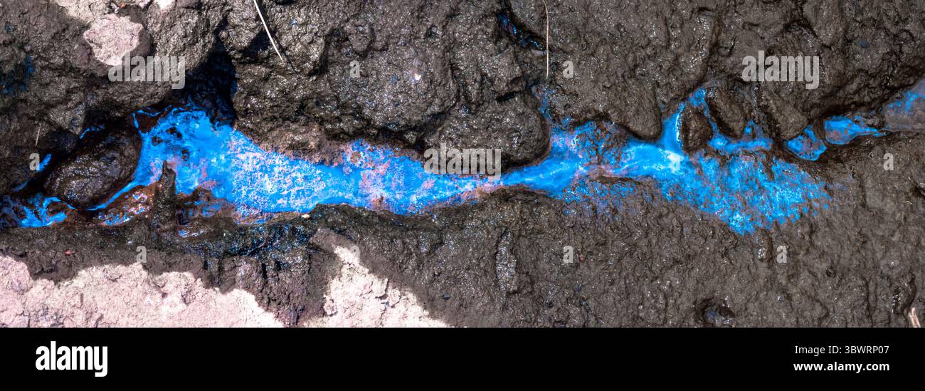 Le liquide bleu électrique s'écoulant à travers un sol fissuré foncé forme un contraste visuel saisissant, mettant en évidence l'impact des problèmes environnementaux Banque D'Images
