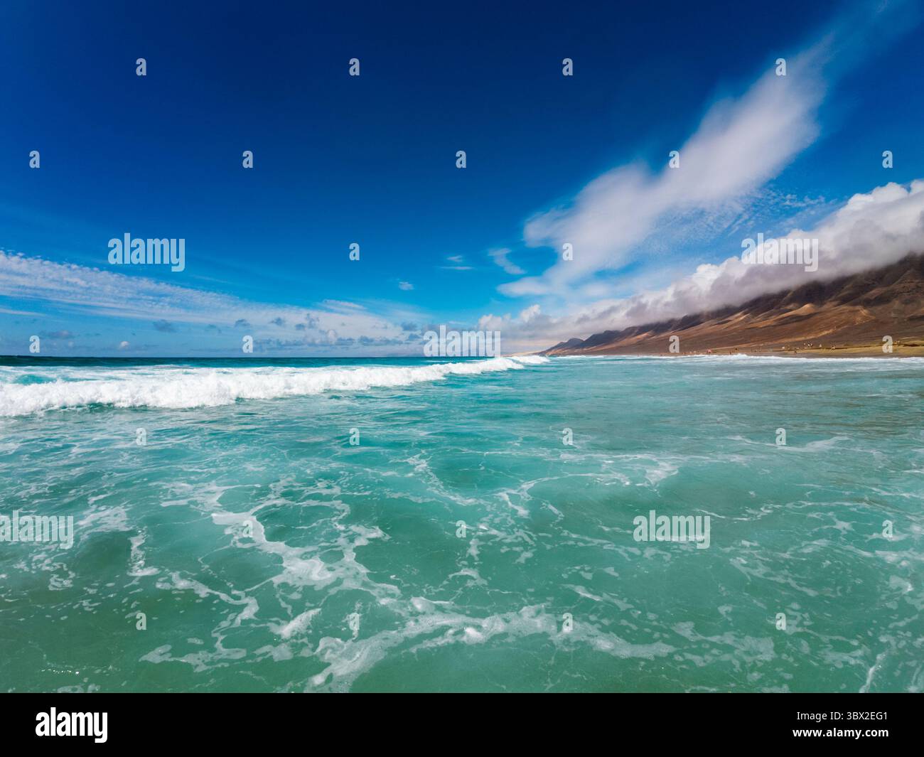 Des vagues turquoises se battent contre la plage de sable de Fuerteventura sous un ciel bleu clair, créant une atmosphère côtière sereine. Banque D'Images
